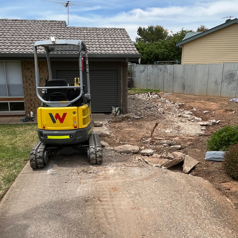 Mini excavator on a driveway beside a front yard strewn with broken concrete