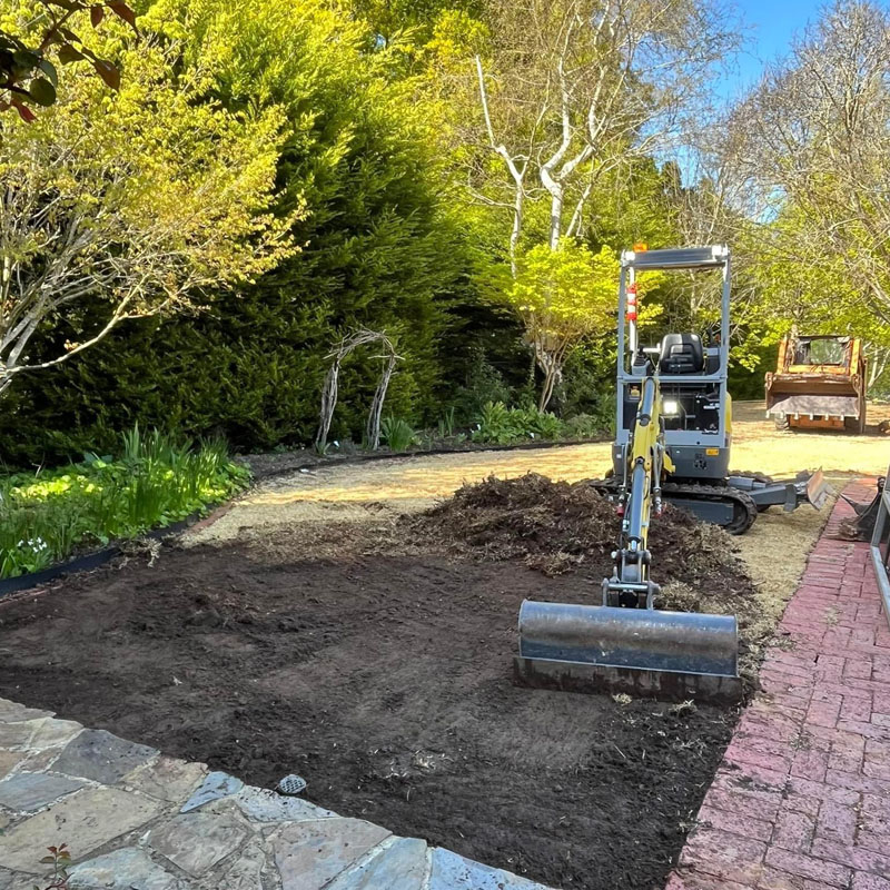 Mini excavator levelling soil in a garden bed beside a brick path