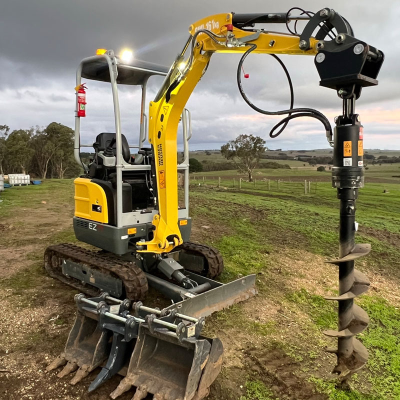 Mini excavator with an auger fitted and spare buckets in front, in a paddock