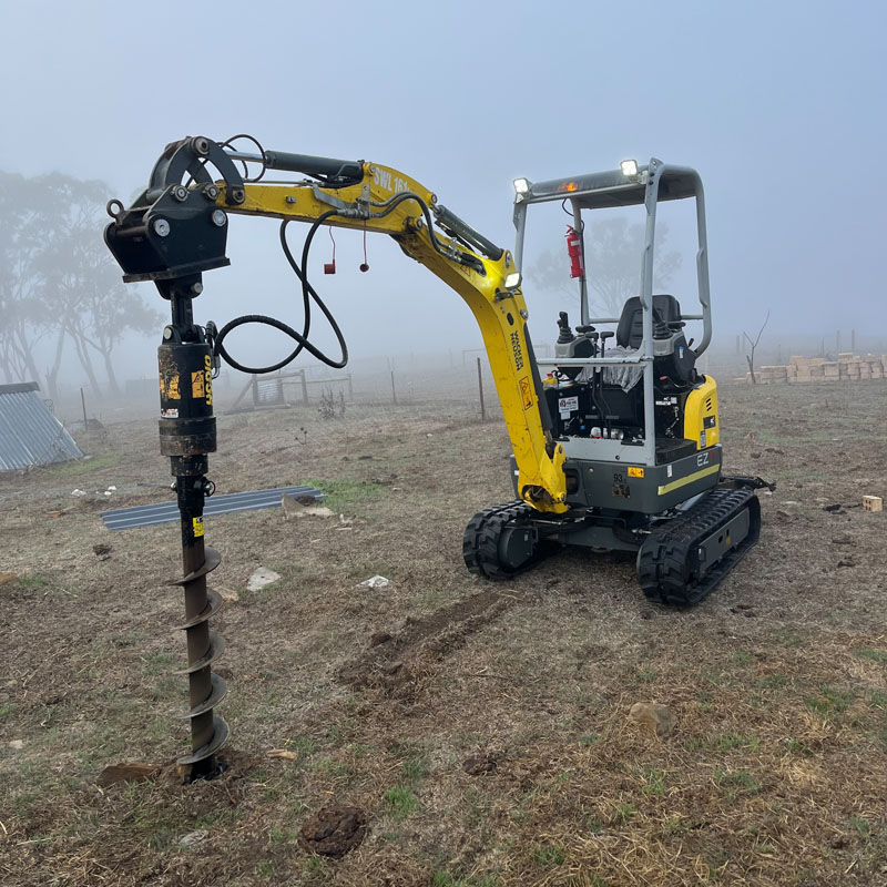 Mini excavator drilling a post hole with an auger in a foggy paddock